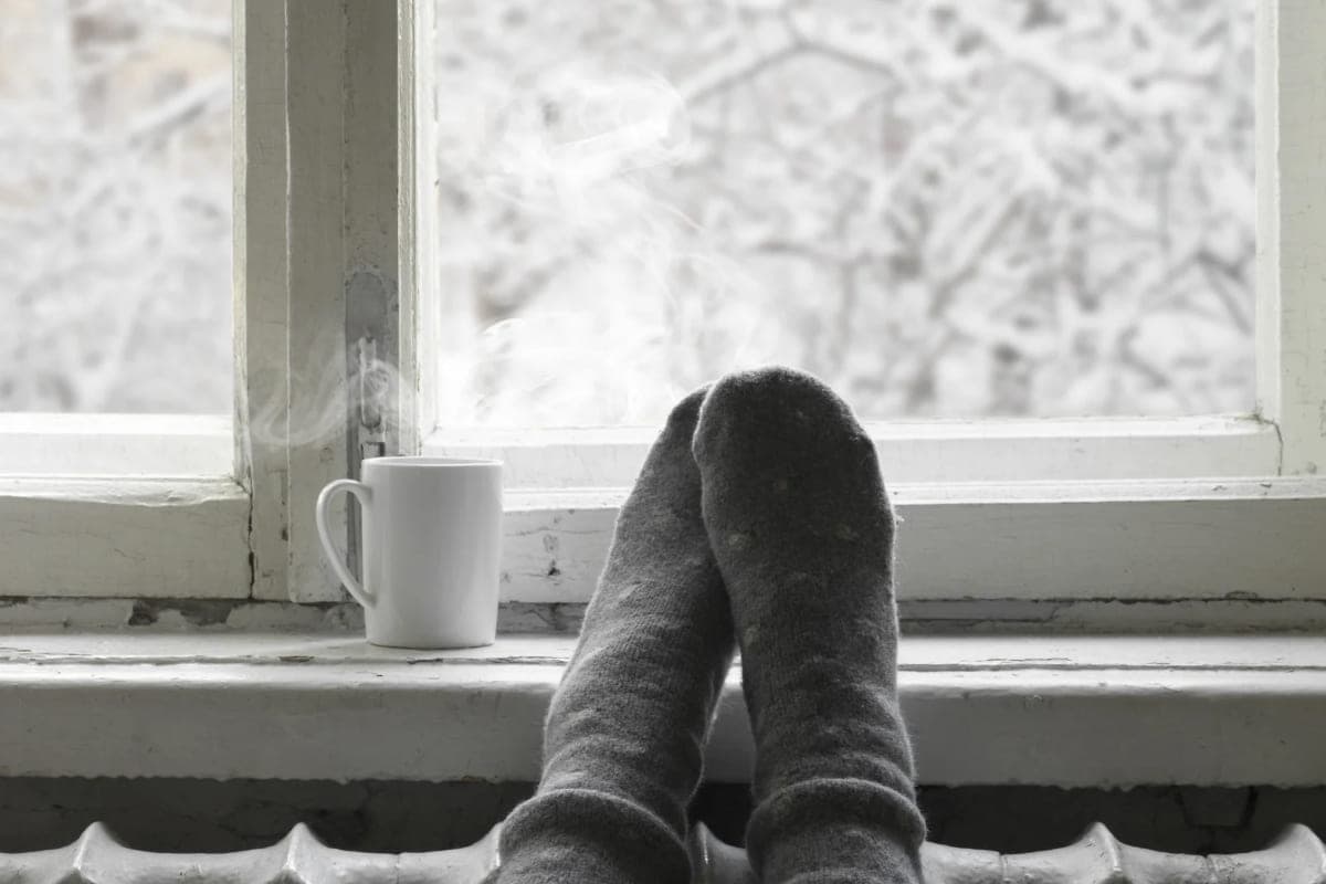 Feet in thick grey wool socks resting on a radiator beneath a window, a steaming mug on the sill, snow-covered branches blurred through the frosted glass — the two conditions winter skin lives between: cold outside and dry heated air indoors.